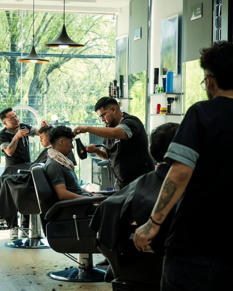 Three barbers working at once in a bright barbershop with tall windows