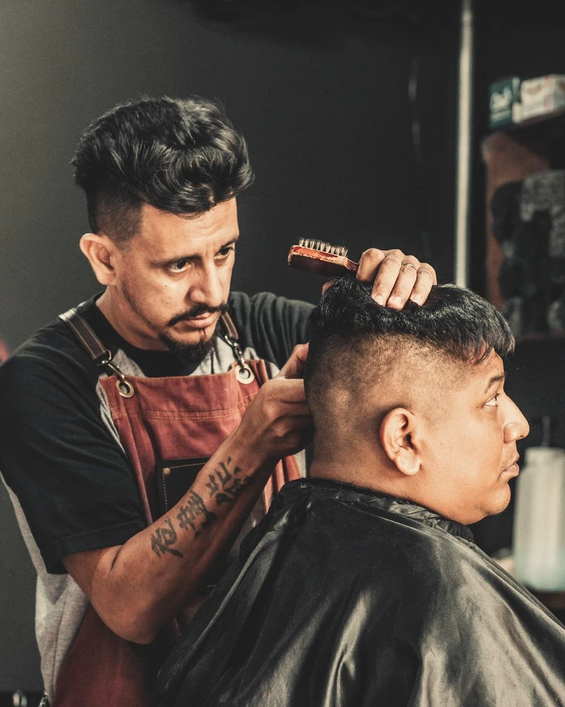 A barber in a leather apron styling a client's hair in the chair