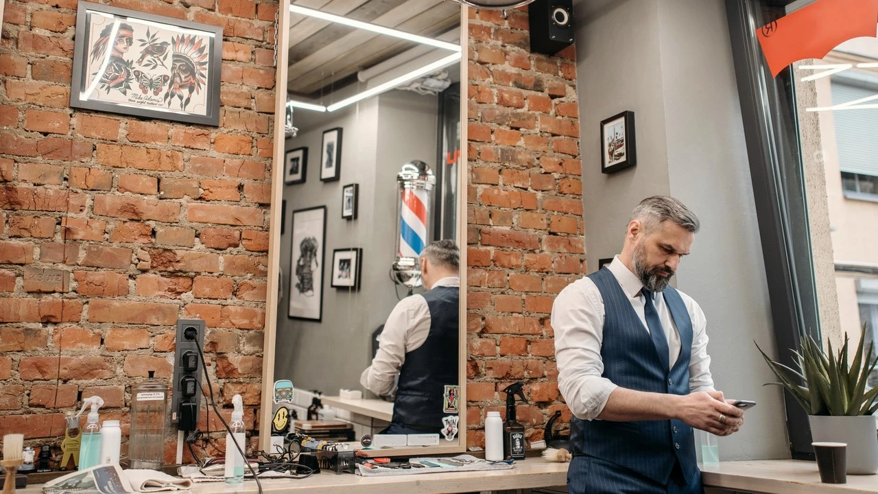 A barber in a waistcoat checks his phone beside the mirror in a barbershop with brick walls